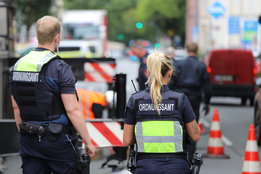 Zwei uniformierte Mitarbeiter des deutschen Ordnungsamts regeln den Verkehr auf einer Baustelle.