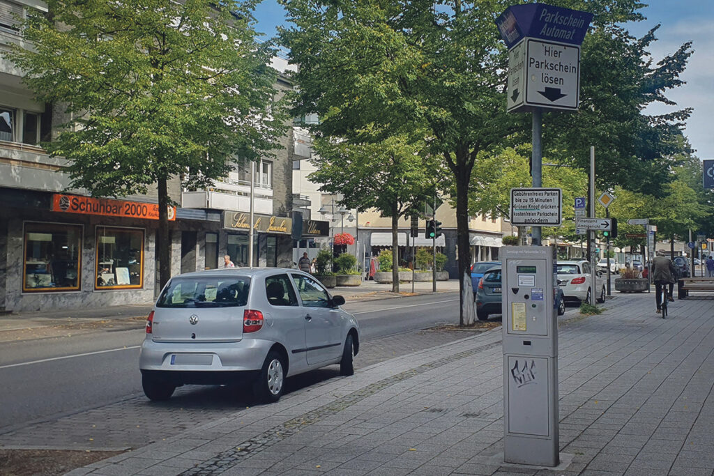 Ein silberner Kleinwagen, der in einer von Geschäften gesäumten Straße geparkt ist; im Vordergrund steht ein Parkautomaten auf dem Bürgersteig.