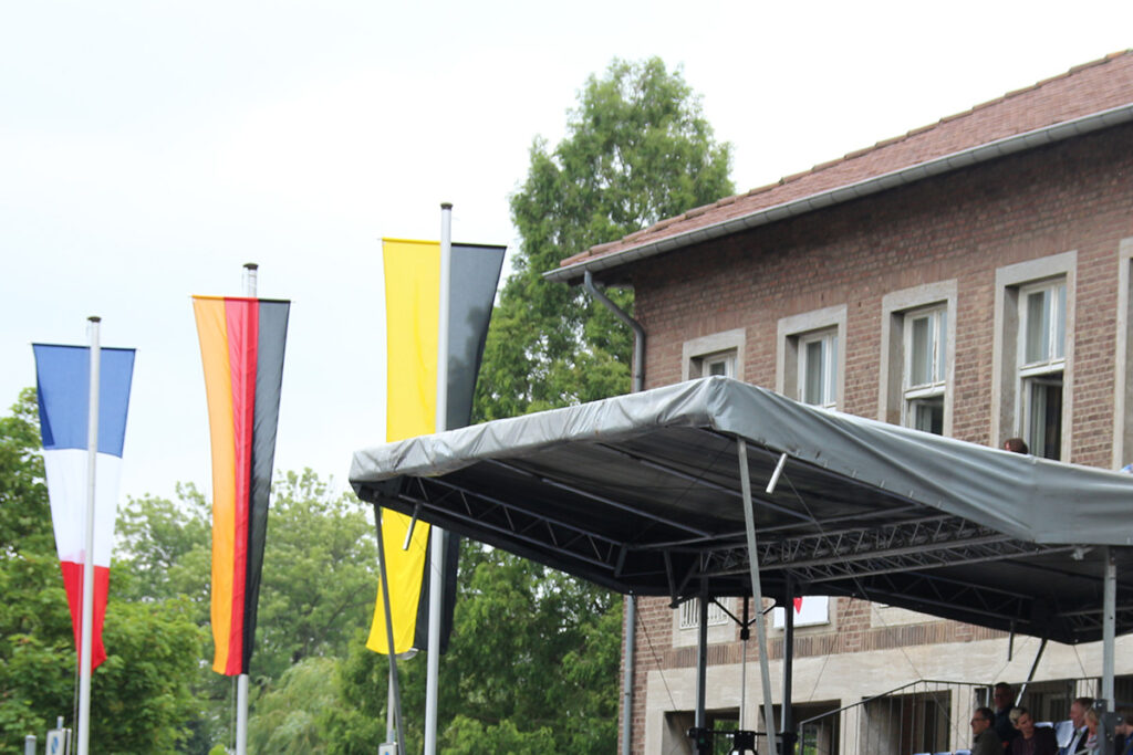 Vor einem Backsteingebäude mit einem Bühnenvordach wehen die Flaggen Frankreichs, Deutschlands und Belgiens.