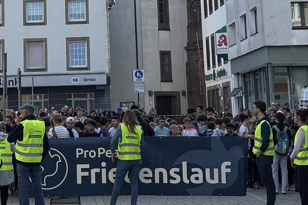 Eine große Menschenmenge steht auf einem Stadtplatz hinter einem Transparent mit der Aufschrift ProPead Friedenslauf.