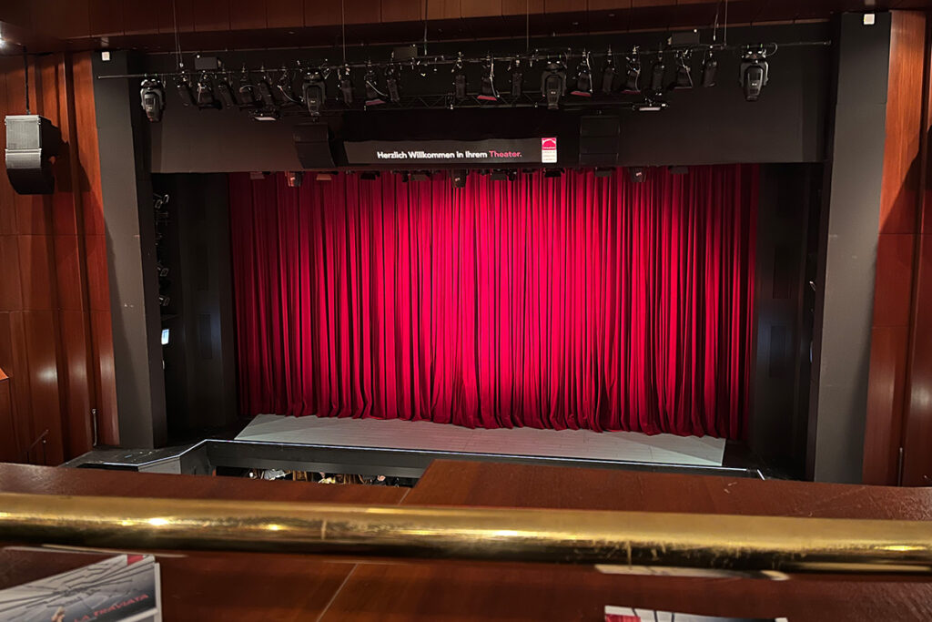 Ein Blick auf eine Theaterbühne mit geschlossenen roten Samtvorhängen und einer digitalen Anzeige darüber, auf der Herzlich Willkommen in Ihrem Theater zu lesen ist.